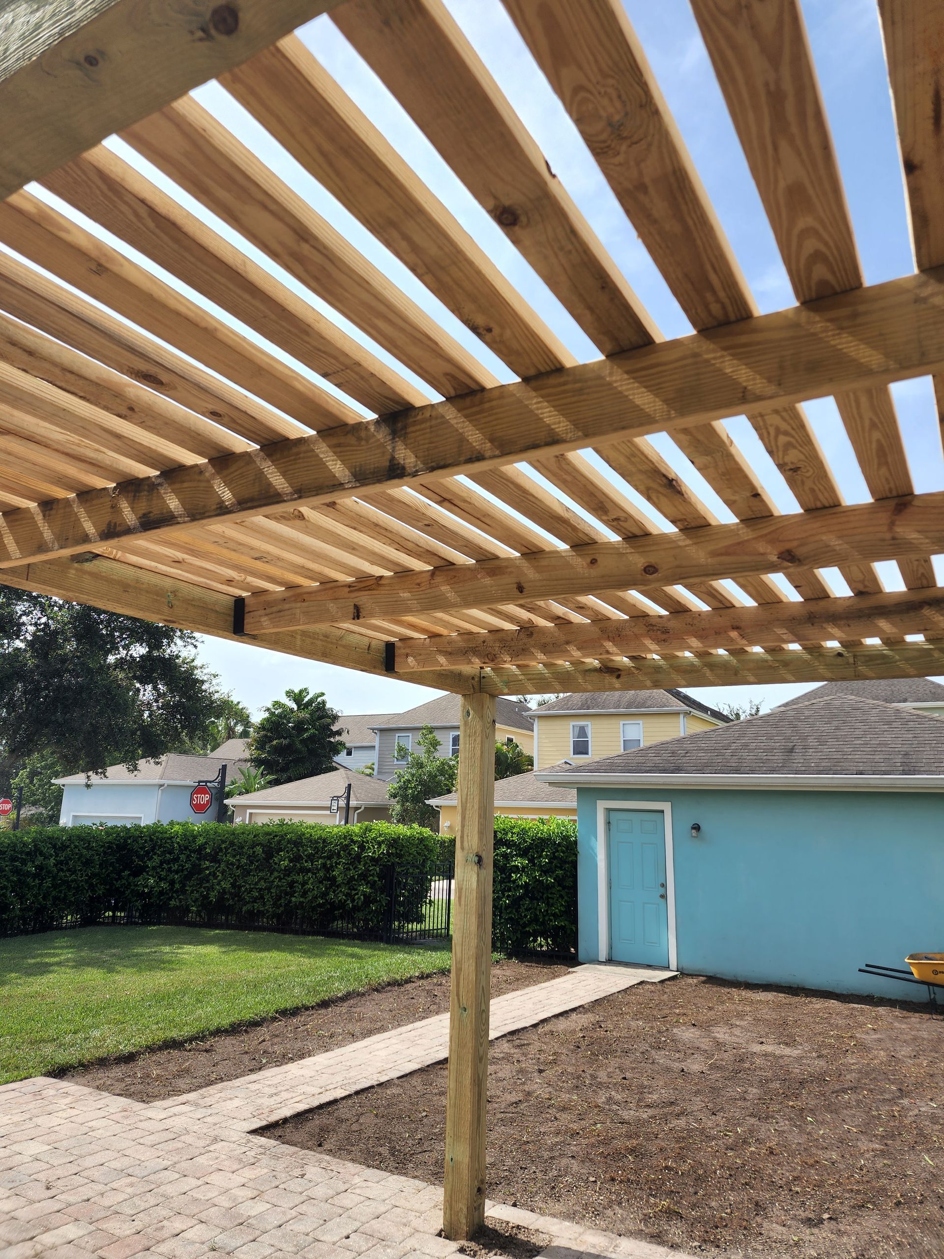 Wooden pergola with horizontal slats, supported by posts, casts shadows on a brick patio. Blue sky peeks through slats.