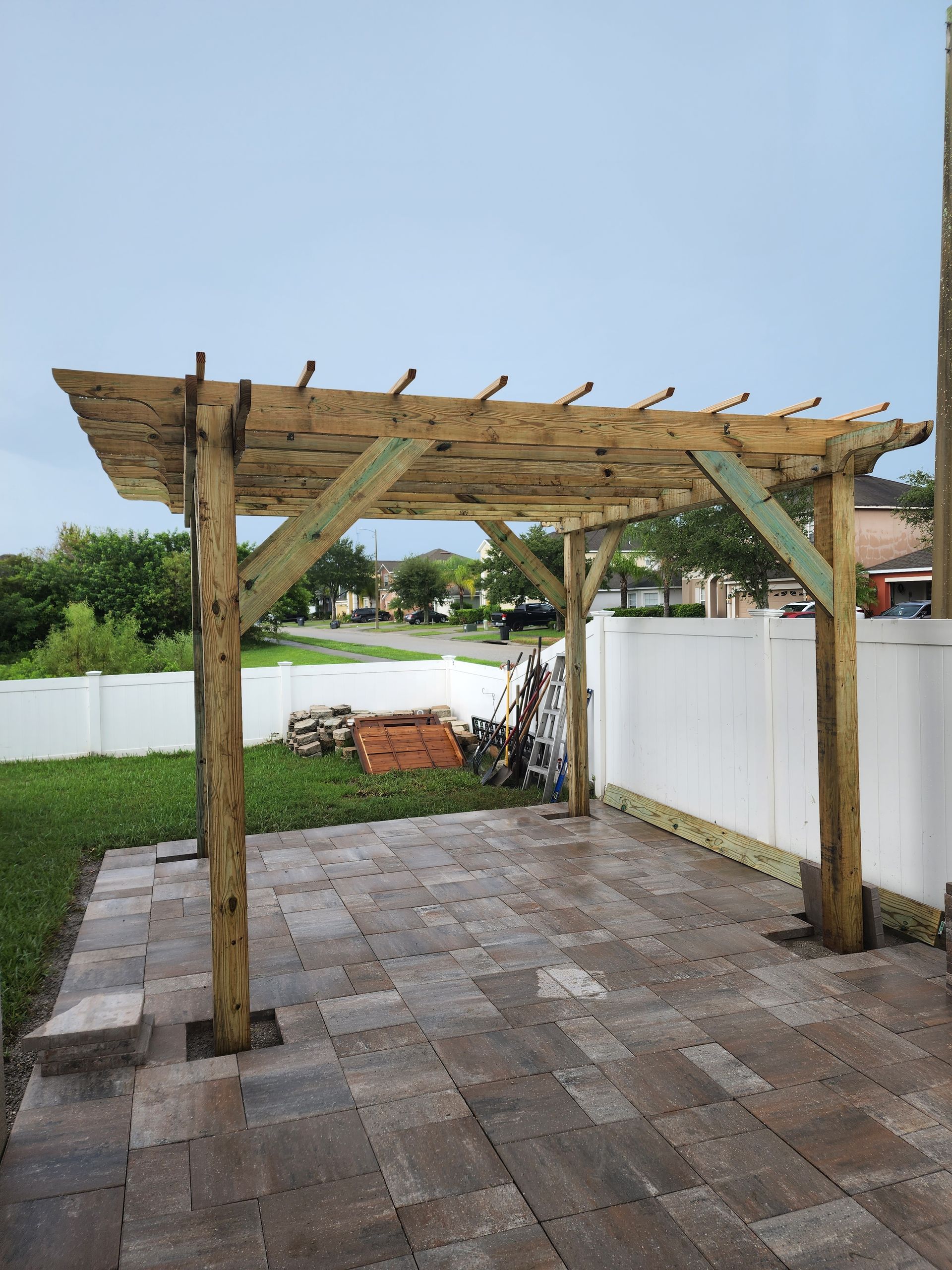 Wooden pergola structure on a paved patio with a white fence and grass in the background under an overcast sky.