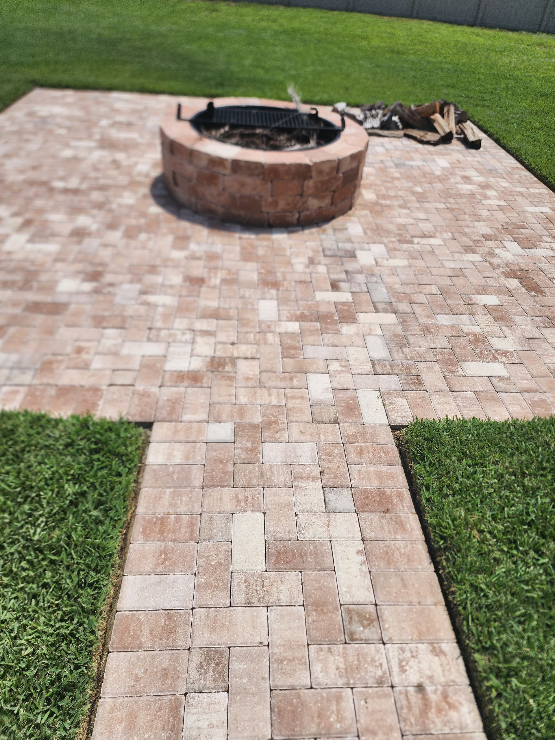 Brick patio with a fire pit, connected by a walkway, surrounded by green grass.