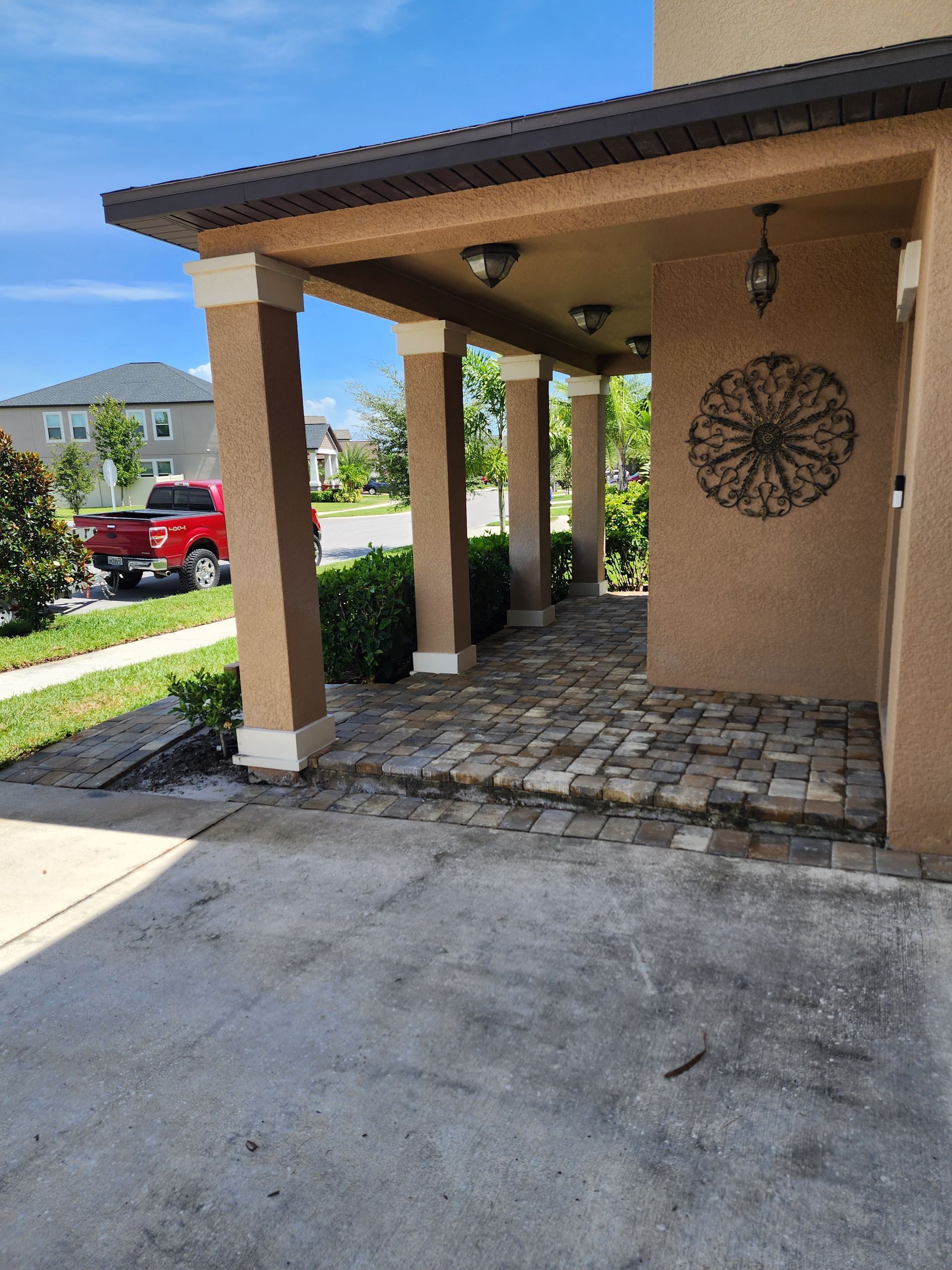 A covered porch with beige pillars and a brick walkway. A red truck is parked in the background on a sunny day.
