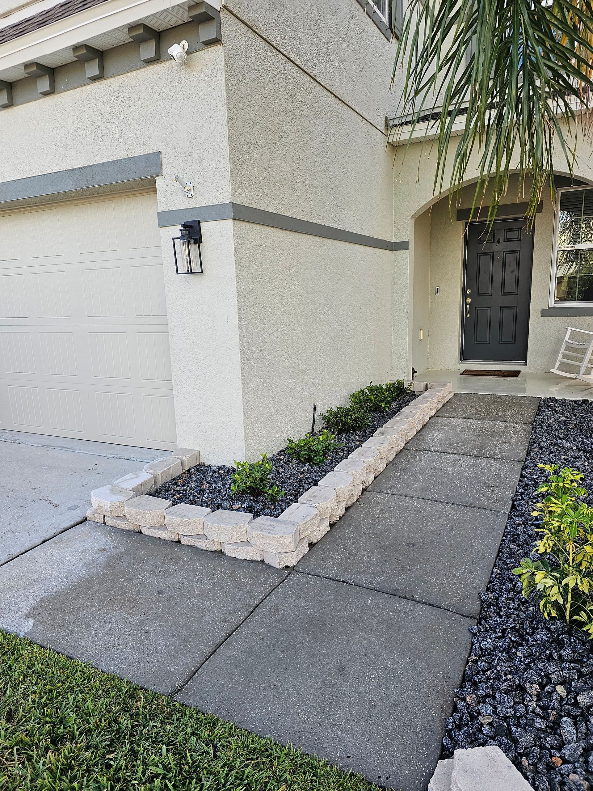 A residential entrance with a raised garden bed lined with light-colored bricks and filled with greenery next to a concrete walkway.