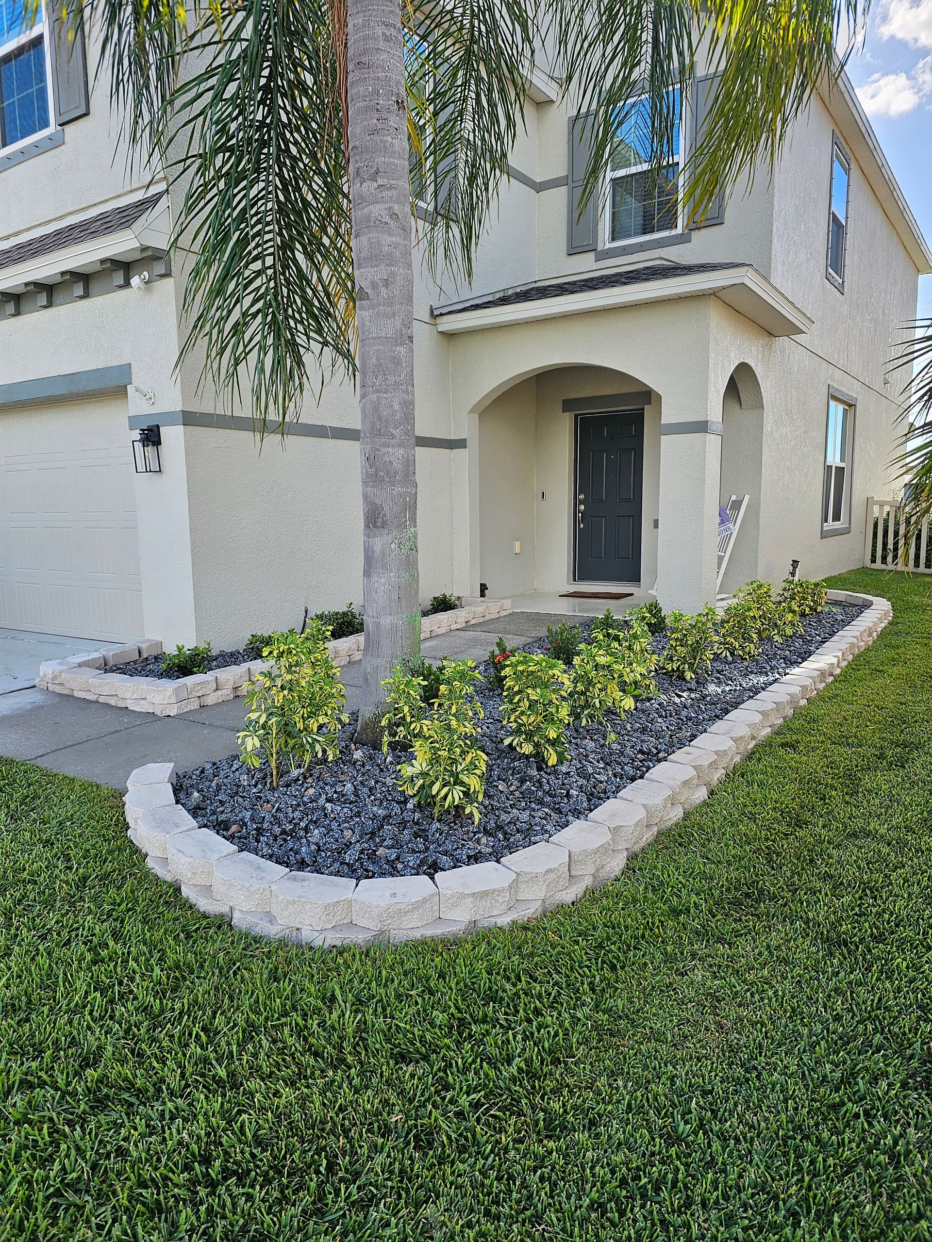 Two-story house with landscaped flowerbed along the walkway, bordered by concrete blocks and filled with black rocks and green and yellow plants.