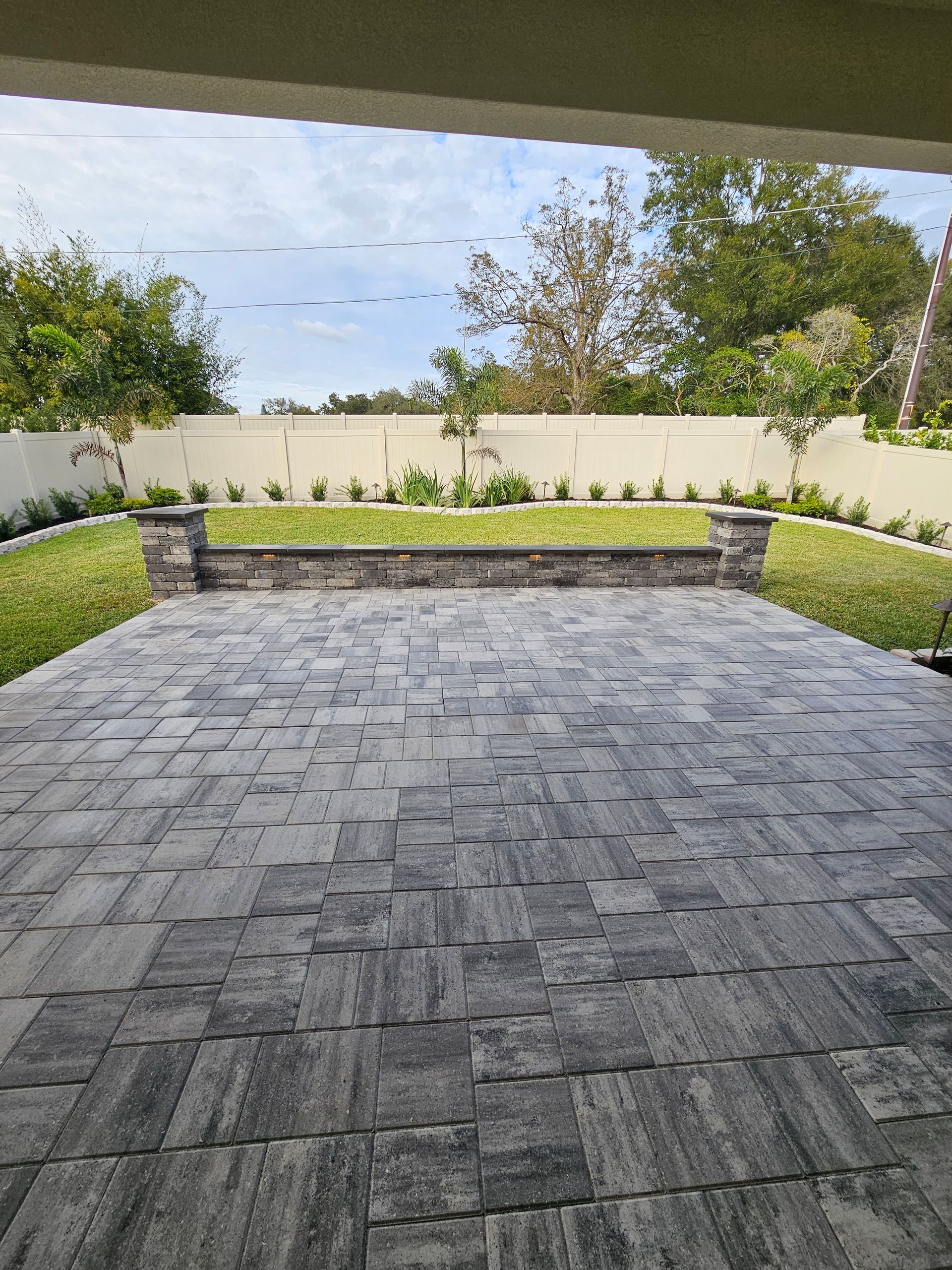 A gray brick patio leads to a raised green lawn with a stone wall and trees against a light blue sky.