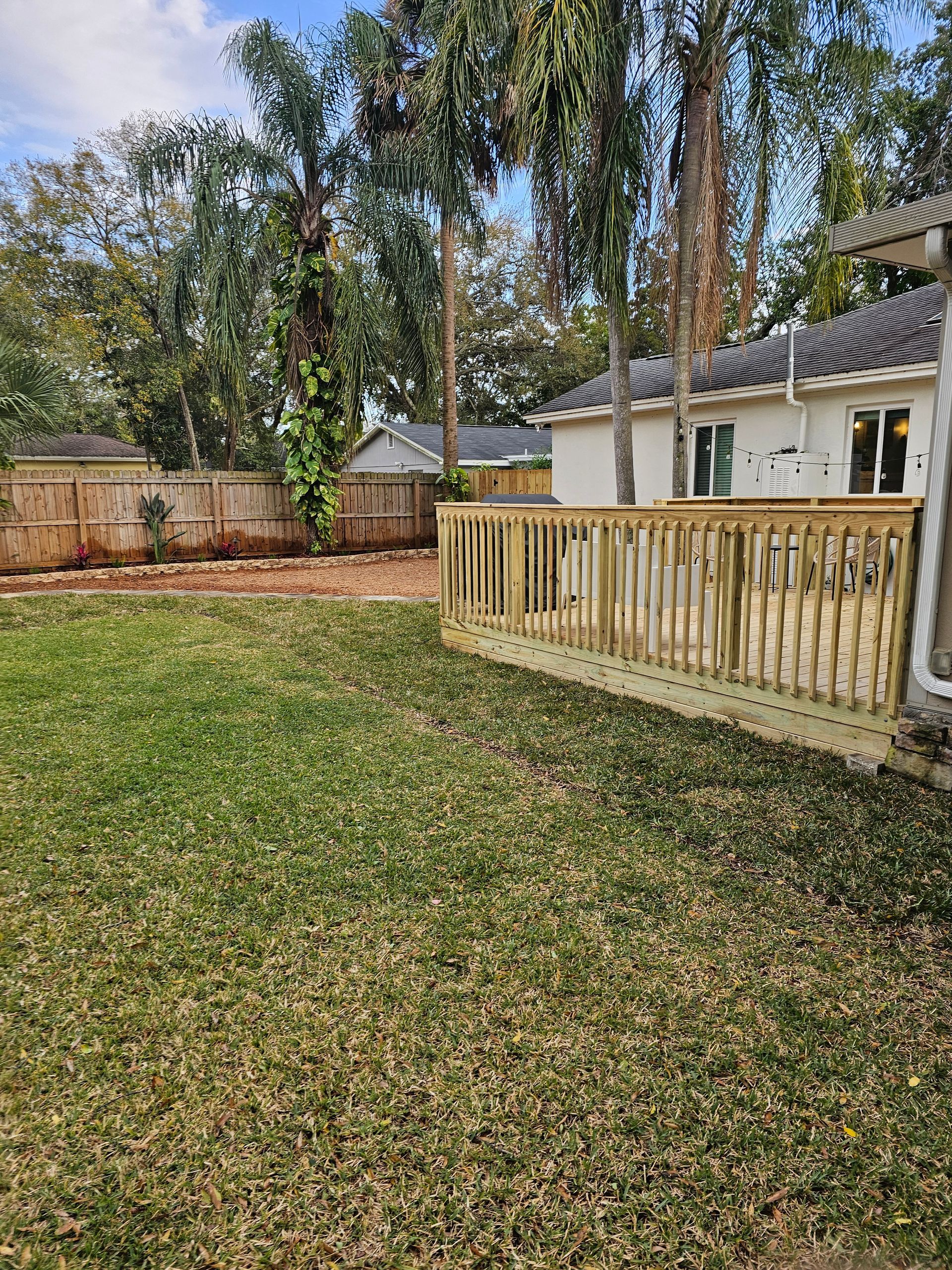 A wooden deck with railings extends from a white house. Green grass and trees surround the deck in a backyard setting.