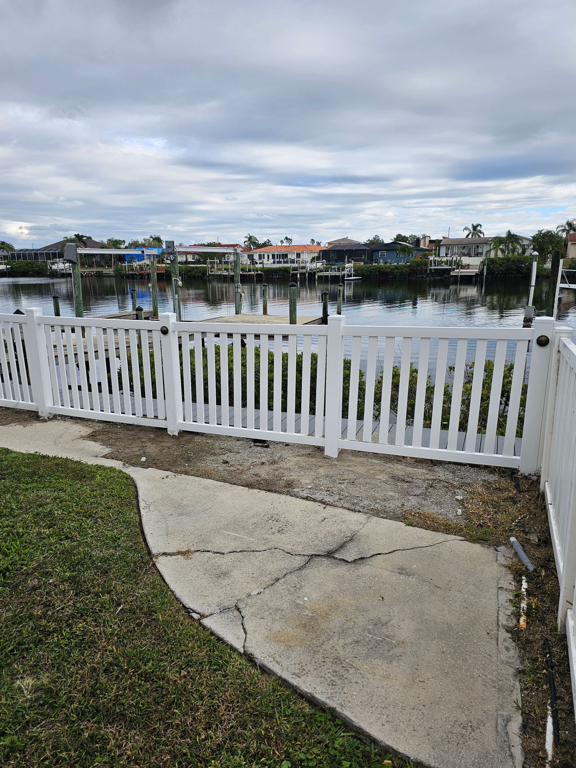 White picket fence bordering a waterway lined with houses, under a cloudy sky. Green grass and a cracked concrete path in the foreground.