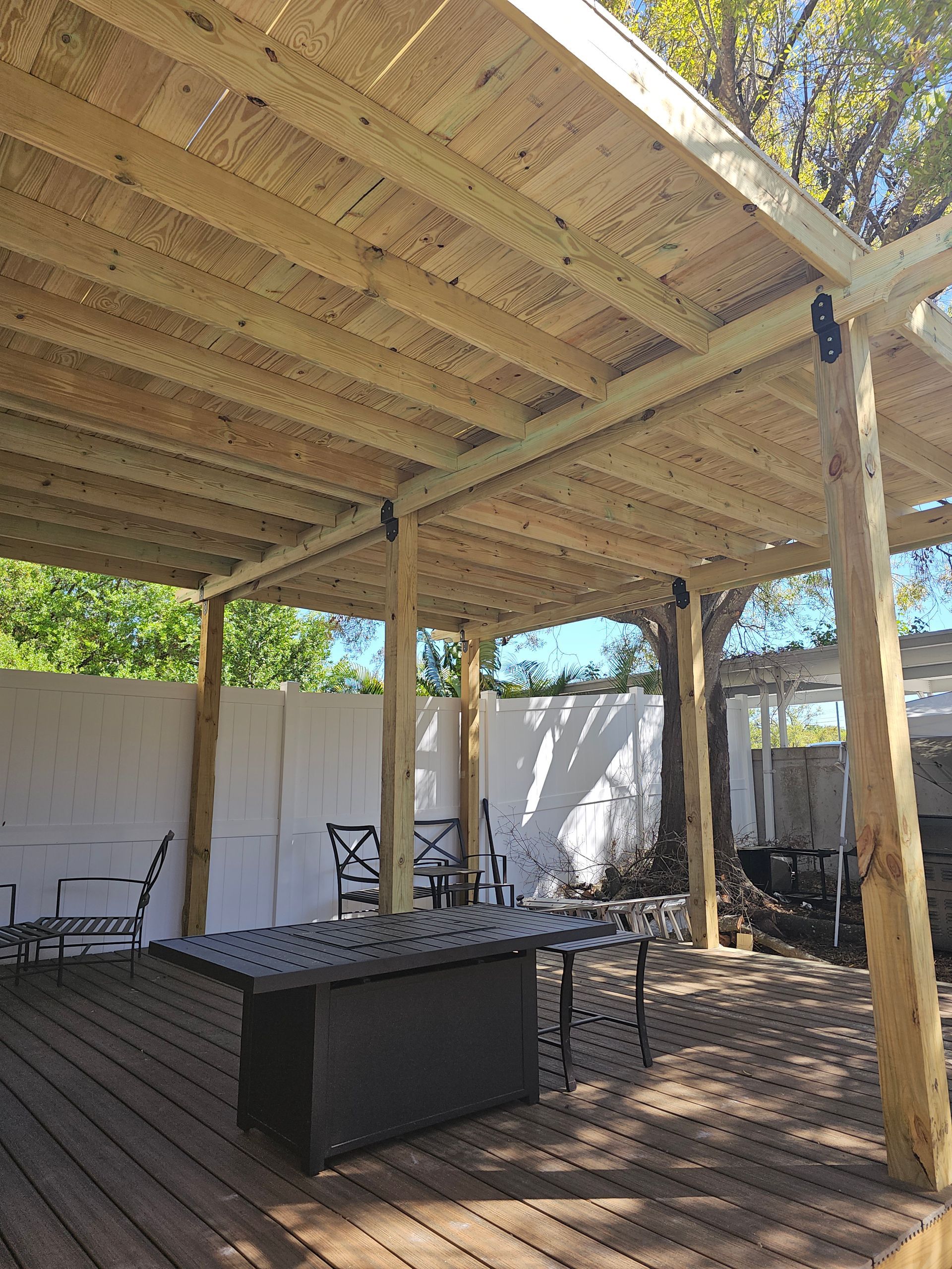 Outdoor wooden patio with a fire table, chairs, and a canopy supported by posts. Sunlight streams through the canopy.