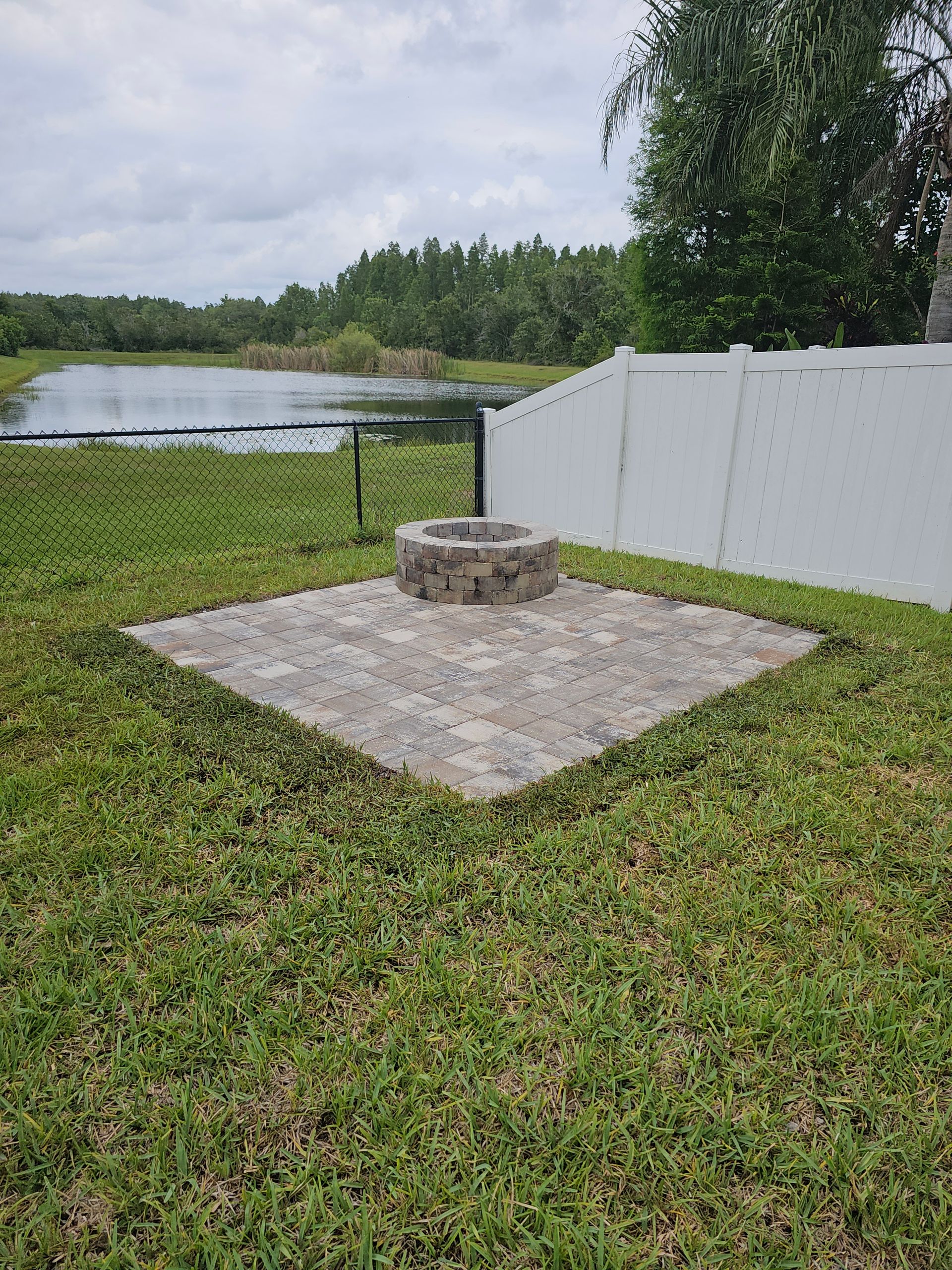 Backyard with a stone fire pit on a paved area surrounded by grass. A lake and trees are visible in the background.