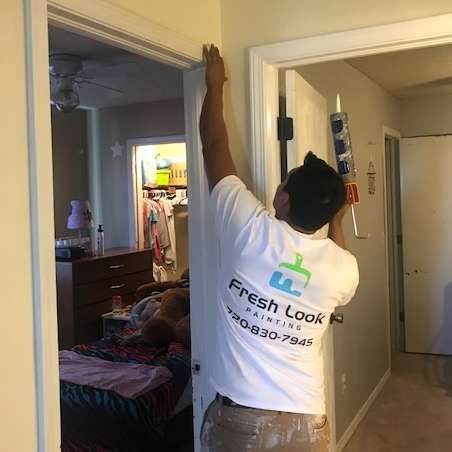 A person caulking a door frame in a hallway. The person wears a white shirt with 