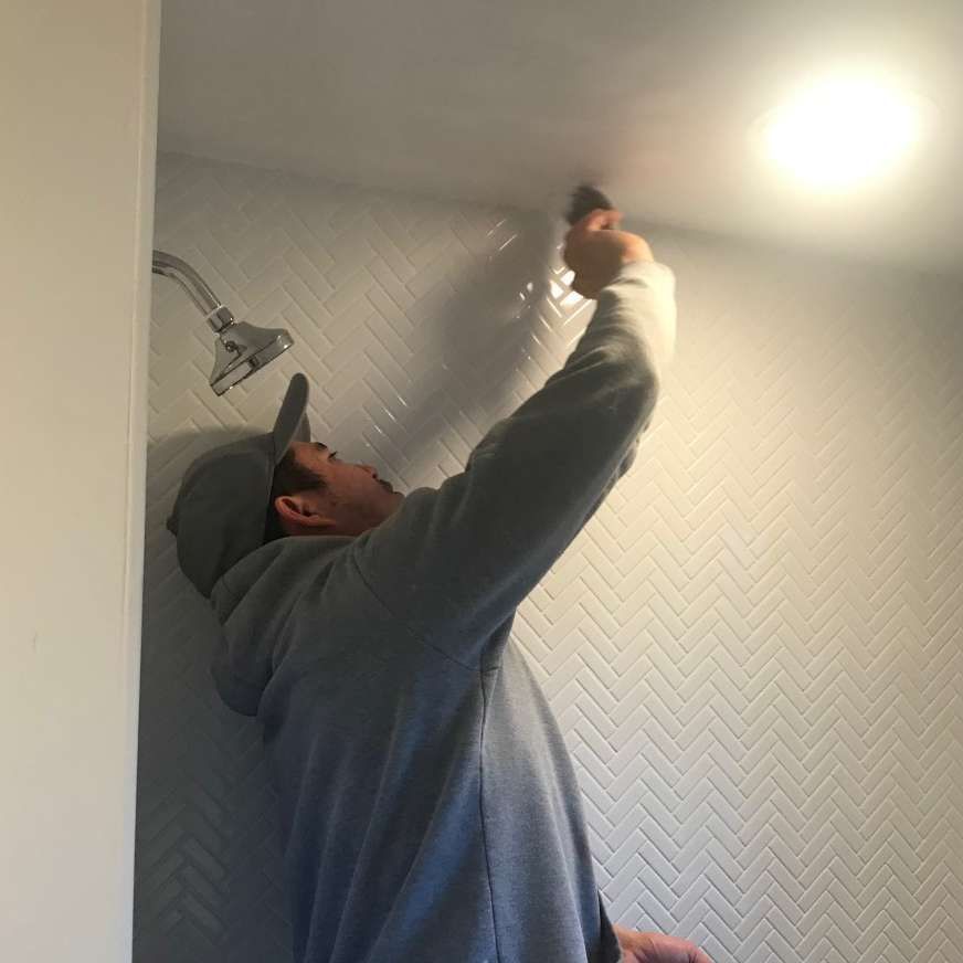 Person installing tile in a shower, using a tool. Light overhead. Herringbone tile pattern.