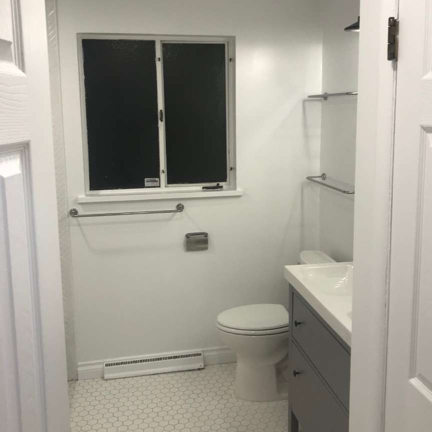 Small, white bathroom with a window, toilet, vanity, and a towel rack.