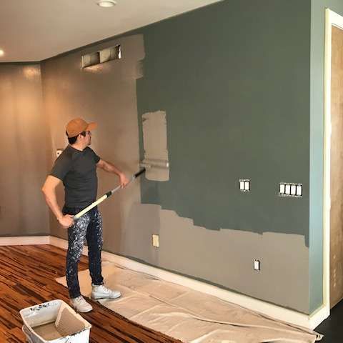 Person paints a wall two-tone green, rolling paint with a roller. Interior room with hardwood floors.