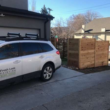 White SUV with roof rack parked beside a garage. A wooden privacy fence is on the right.