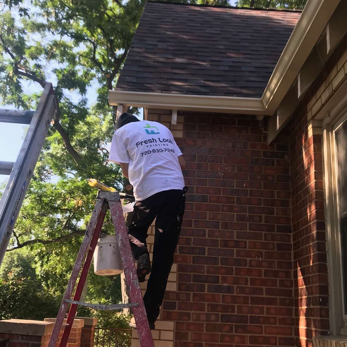 Person on a ladder painting a brick wall, holding a paint can.