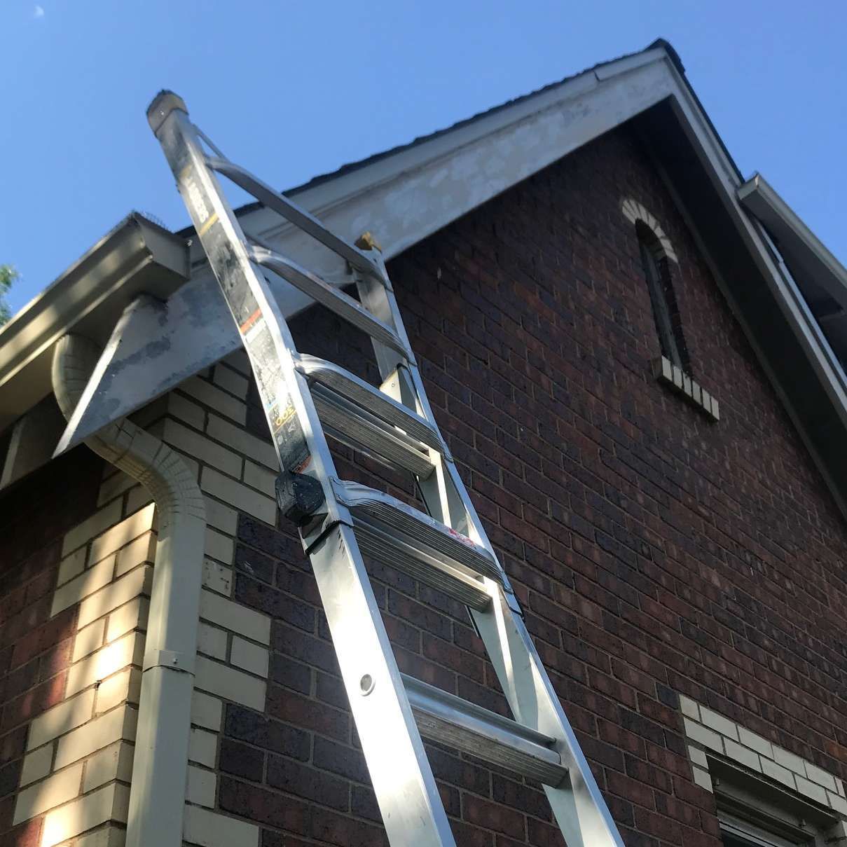 Ladder leaning against a brick house with a dark roof and a clear blue sky.