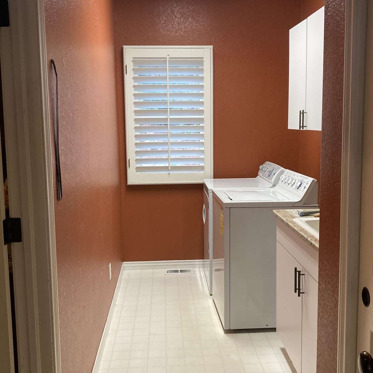 Laundry room with white washer and dryer, window with shutters, and white cabinets against rust-colored walls.