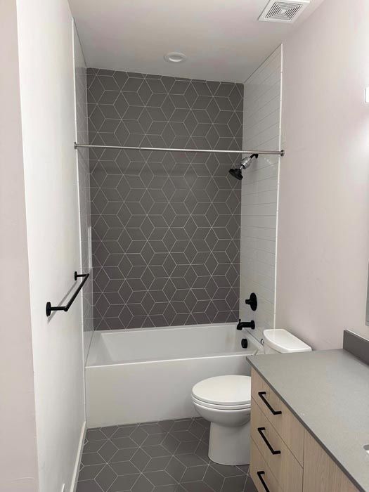 Bathroom with a white tub, toilet, and light-colored vanity. Gray hexagon-patterned tile on the shower wall.