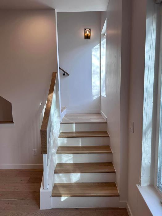 Staircase with light wood treads, white risers and walls, natural light from a window. Wall-mounted light.