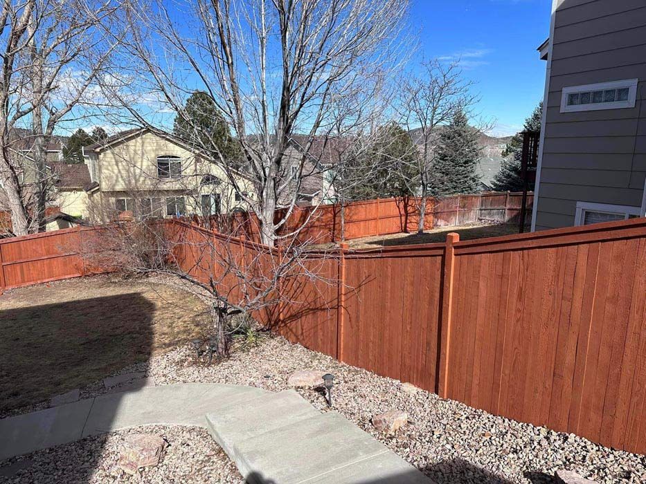 Red wooden fence in a yard with a concrete walkway, gravel, and bare trees under a blue sky.