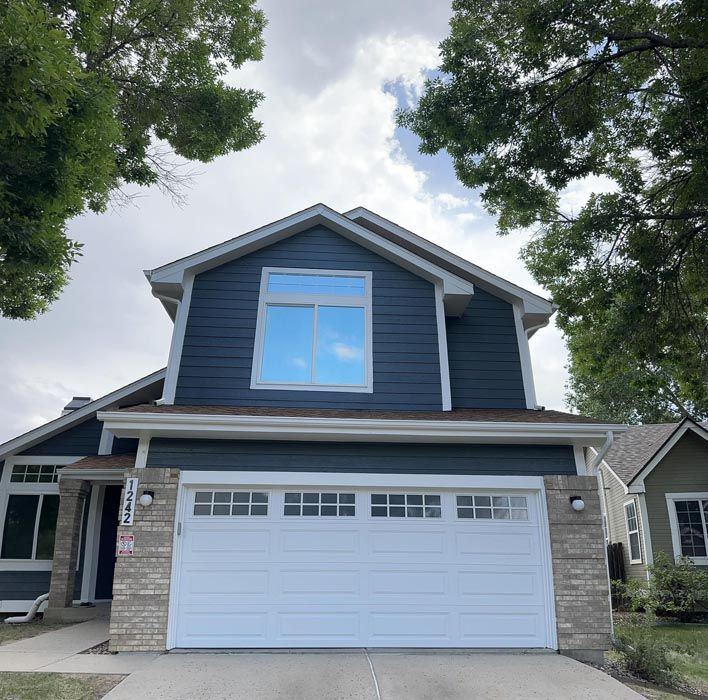 Two-story blue house with white garage door and large window, cloudy sky above.