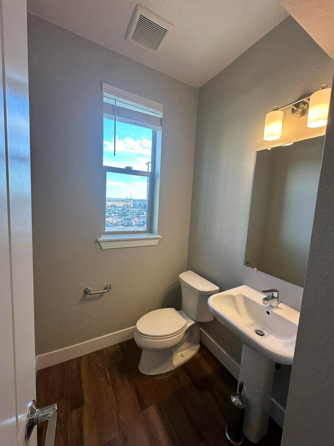 Bathroom with toilet, sink, mirror, and window with an exterior view. Brown wood floor, light gray walls.