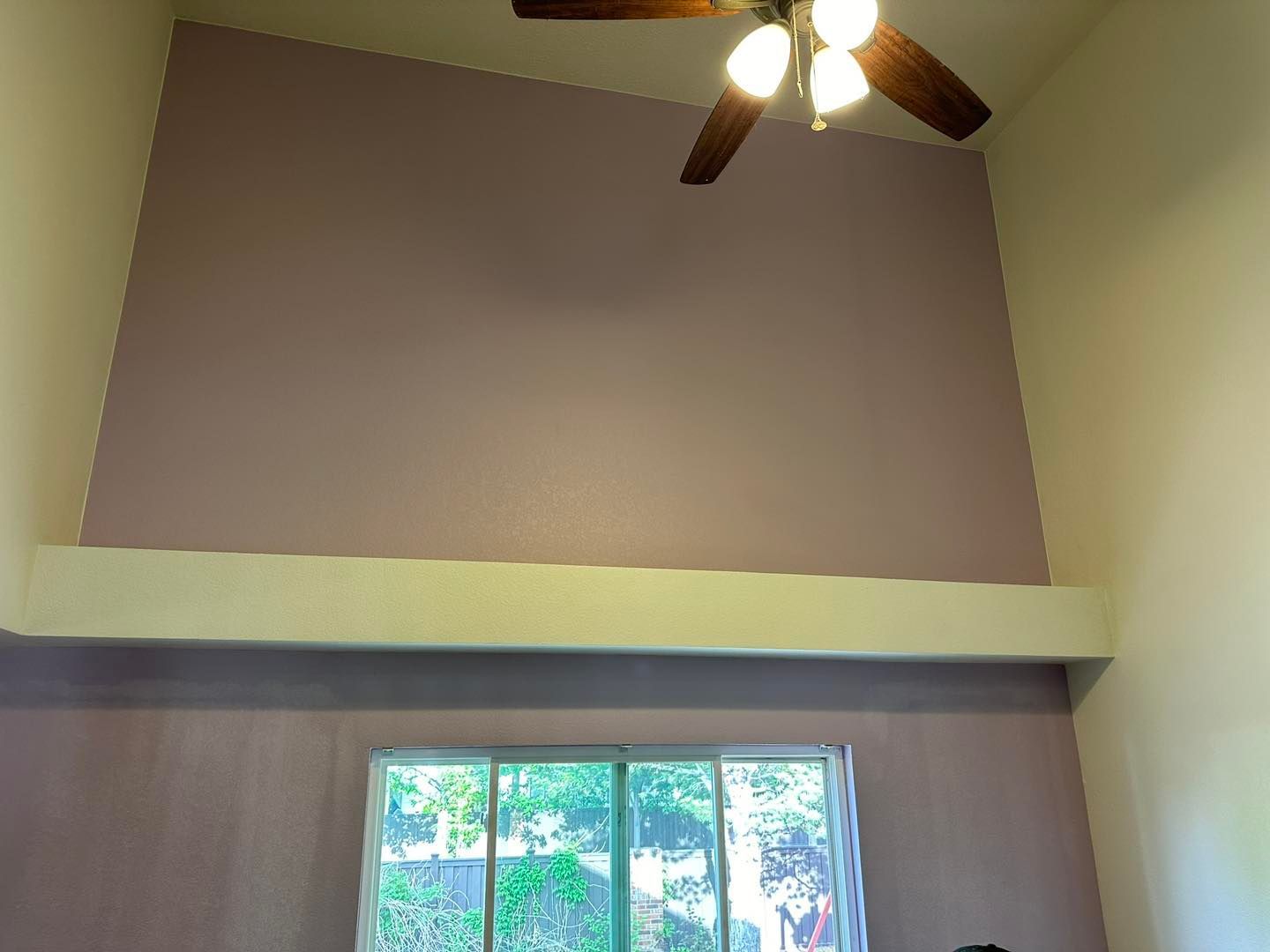 A room with a high ceiling, painted in lavender and cream, has a ceiling fan and a window.