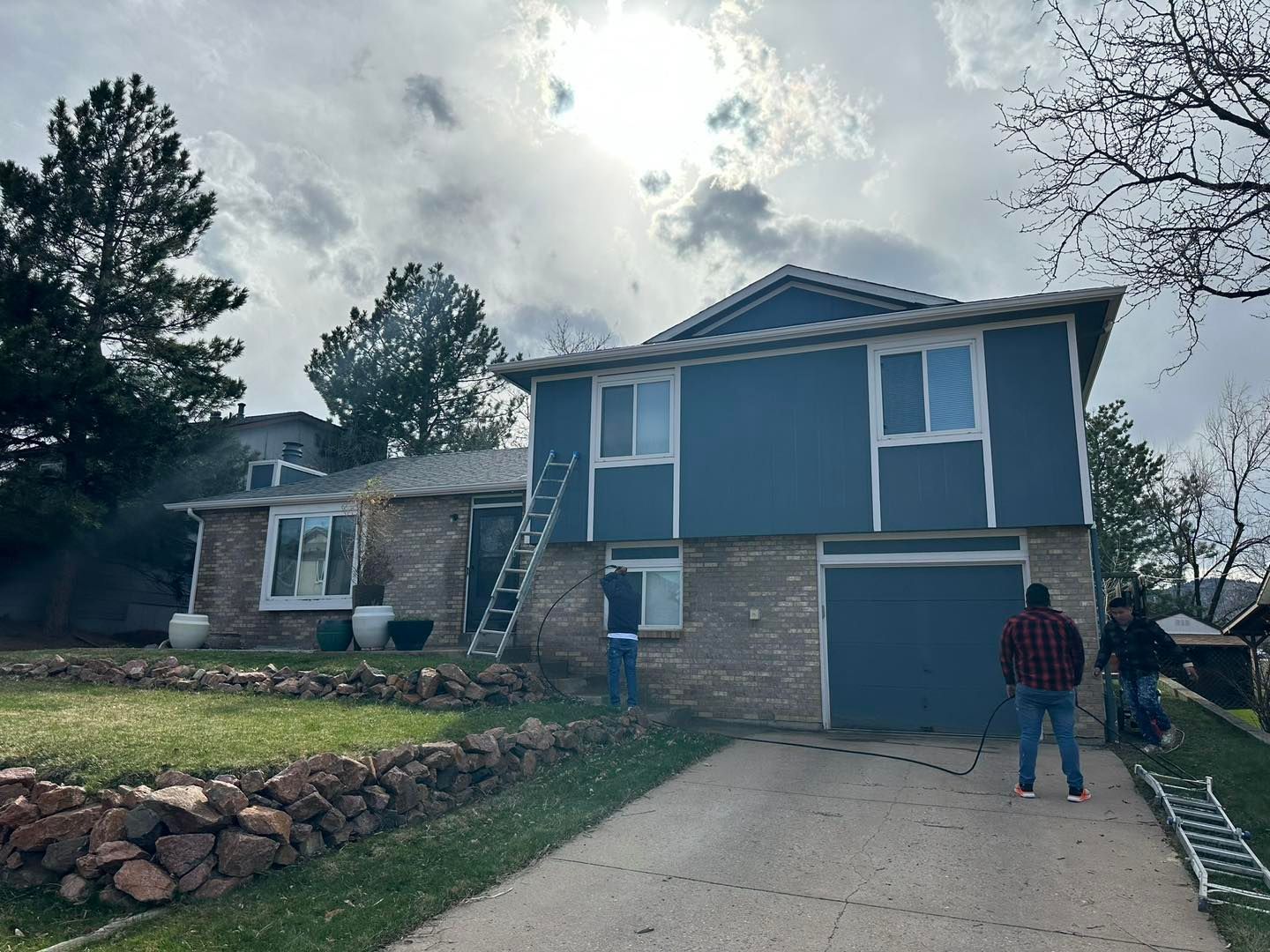 House being painted blue; workers with ladder, cloudy sky, brick facade, grass, driveway.