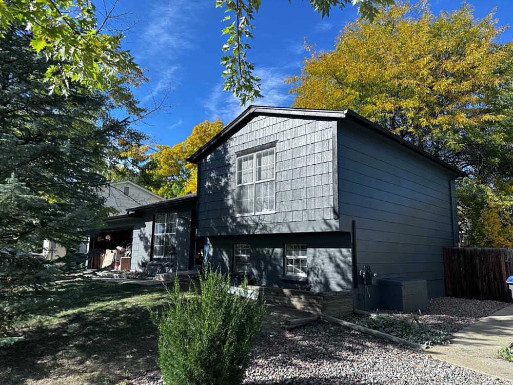 Two-story gray house with shingle siding, trees, and blue sky.