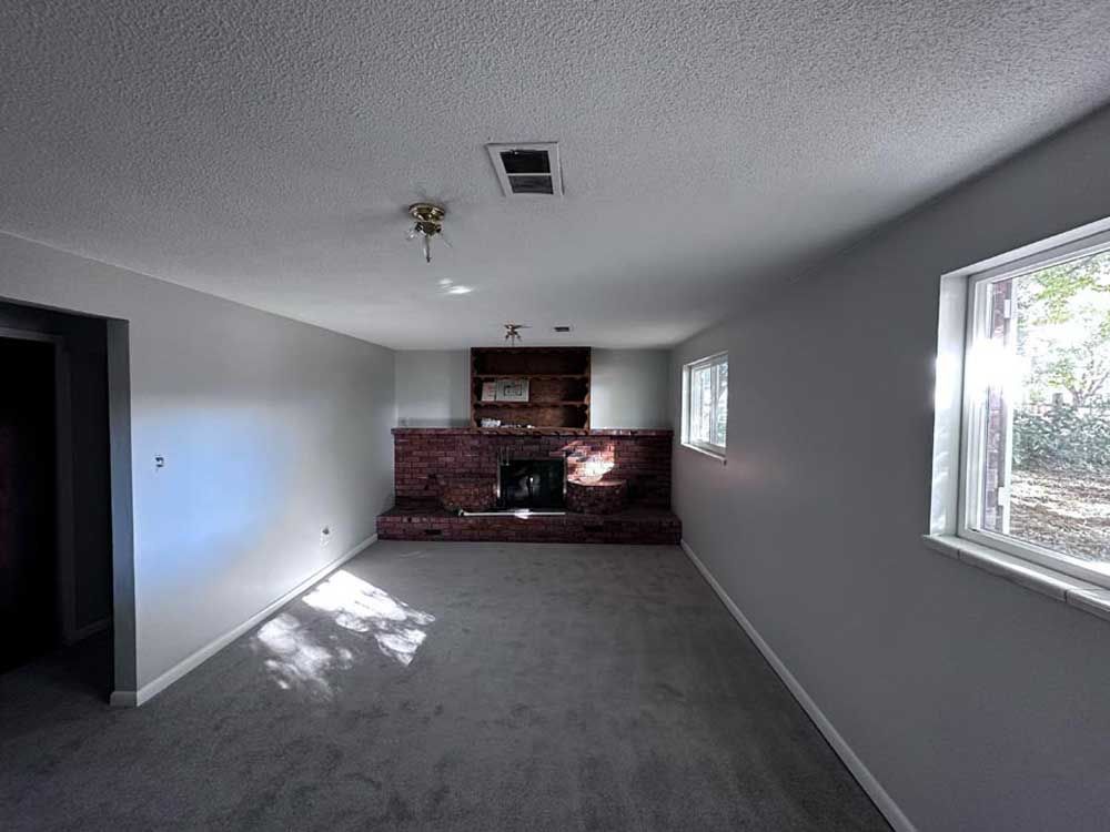 Empty room with gray walls, carpet, and a brick fireplace. Sunlight streams through windows.