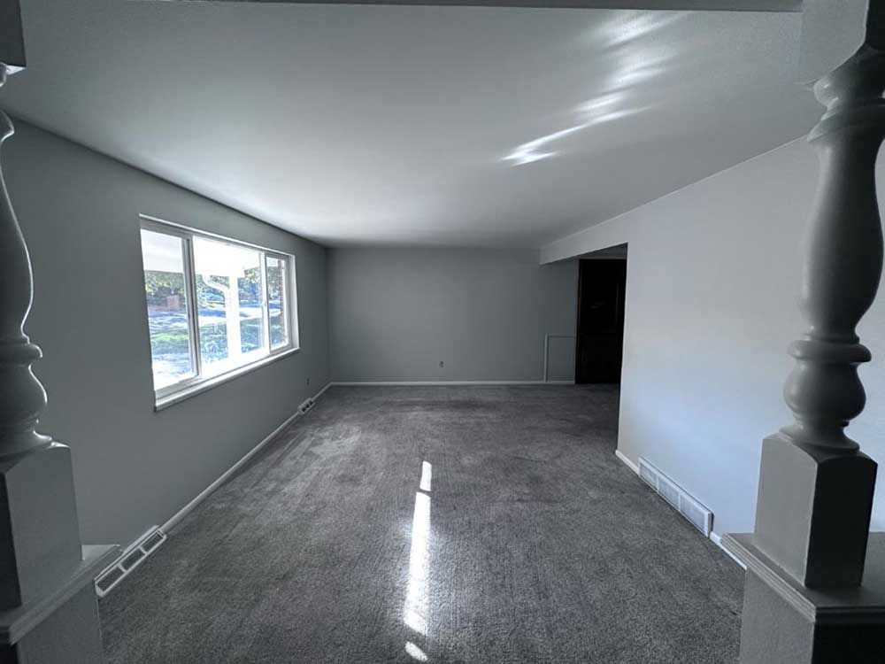 Empty living room with gray carpet and walls; sunlight streams in.