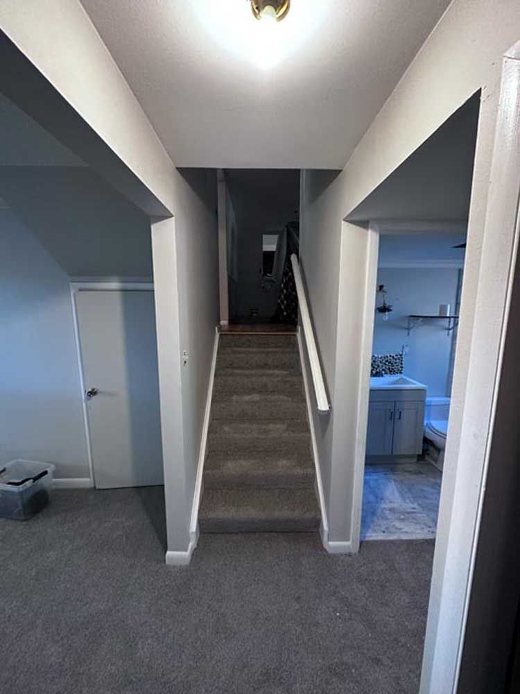 Staircase with gray carpet leading to an upstairs area, flanked by white walls and doorways; a light fixture is centered above.