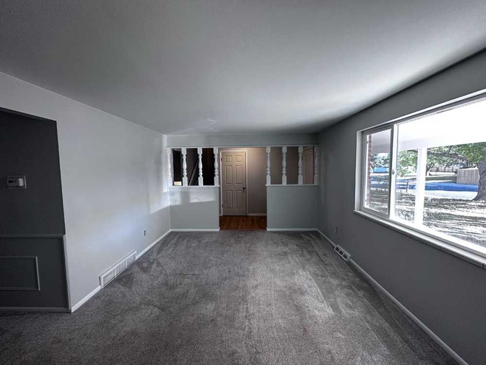Empty living room with gray carpet, walls, and trim. Two windows. White door visible.