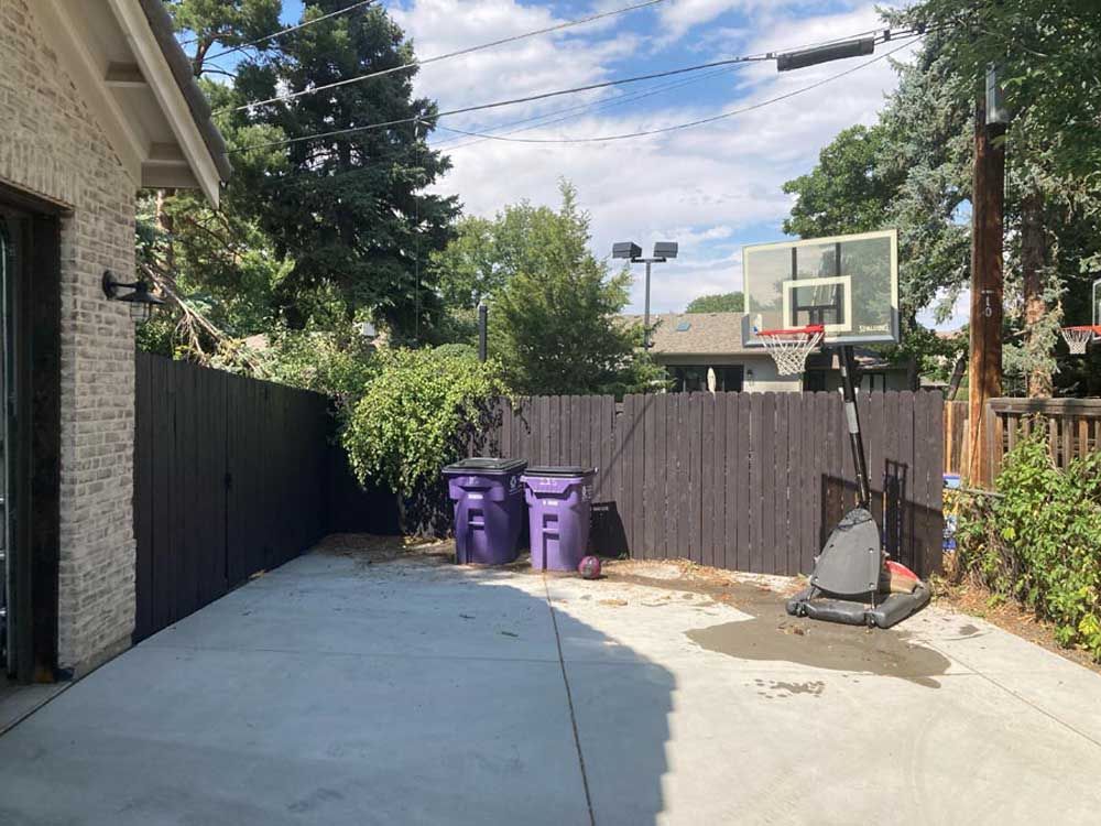 Backyard with dark fence, basketball hoop, purple bins, and a concrete patio.