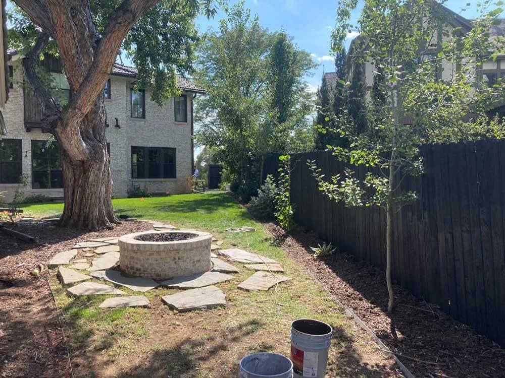 Backyard with fire pit, tree, grass, and dark wooden fence. A light-colored house is visible.