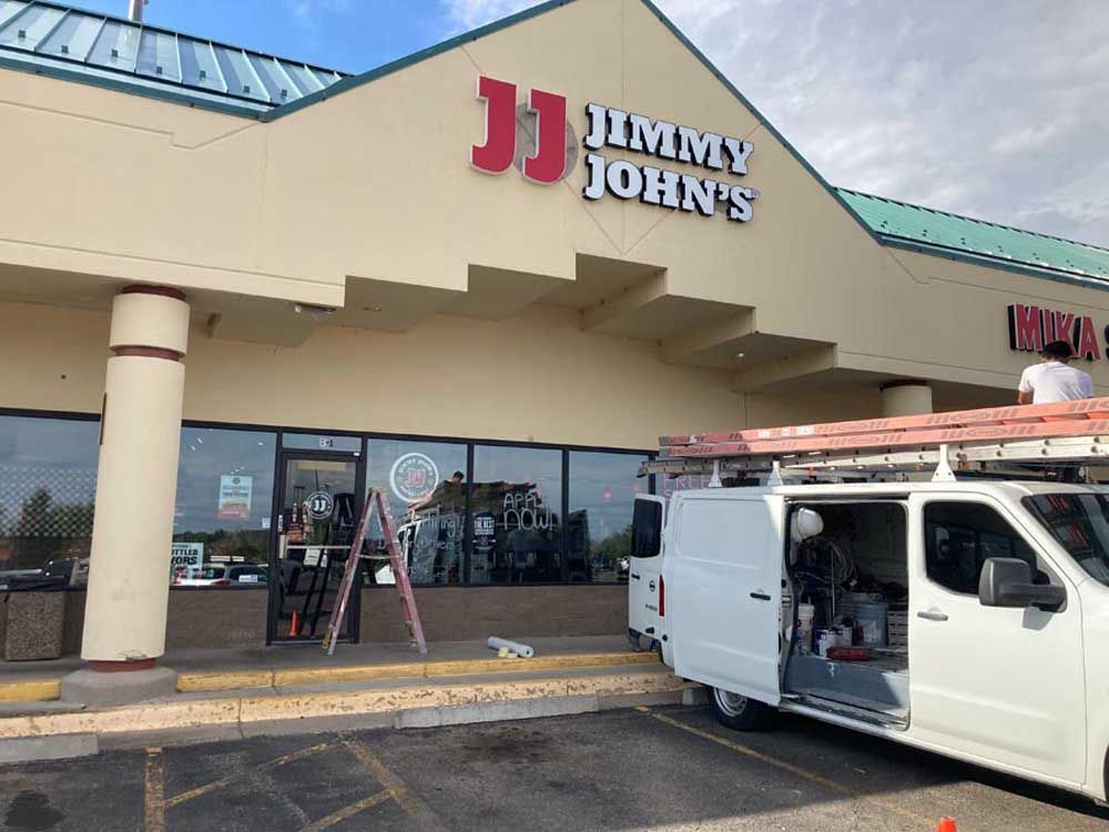 Jimmy John's storefront with workers and van; red and white sign, tan building, ladder on van.
