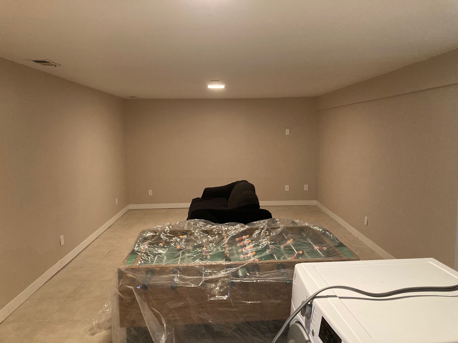 Empty beige-walled room with a covered pool table and a dark couch; a washing machine is in the foreground.