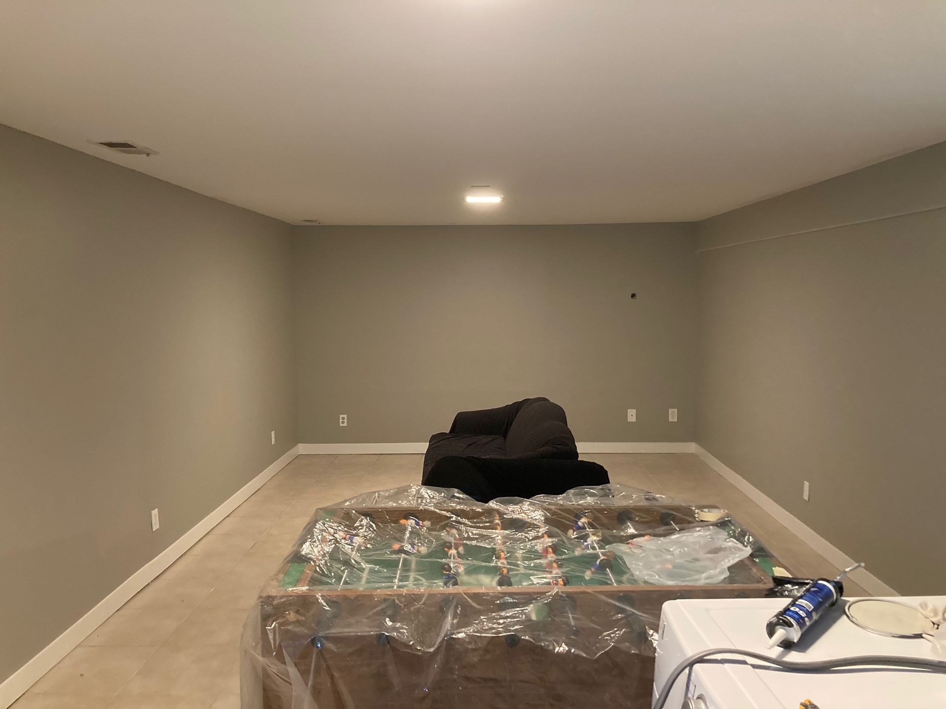Empty room with gray walls and a covered foosball table. Dark couch in the background.