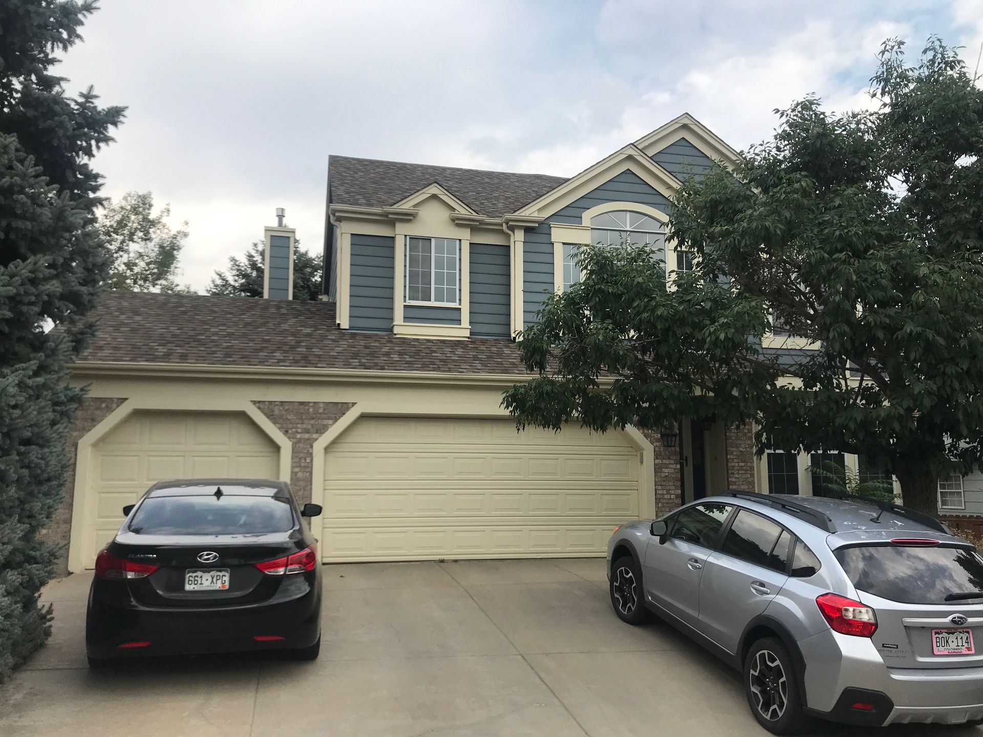 Two-story house with blue siding, two garage doors, two cars in driveway, trees on either side.