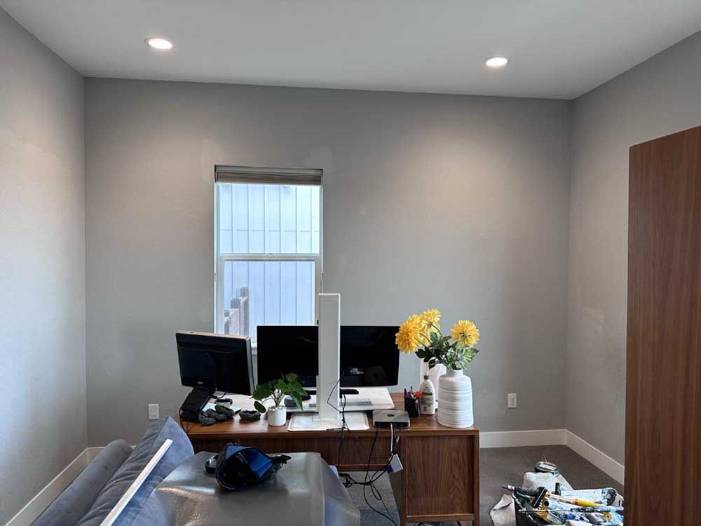 Office space with monitors, desk, window, and cabinet. Gray walls, yellow flowers, and a cap on the desk.