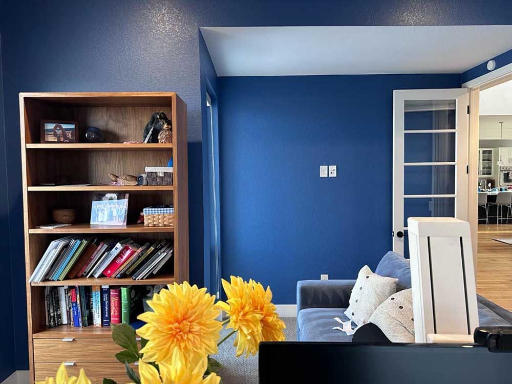 Blue-painted room with wooden bookshelf and sofa, French door to kitchen, yellow flowers in foreground.