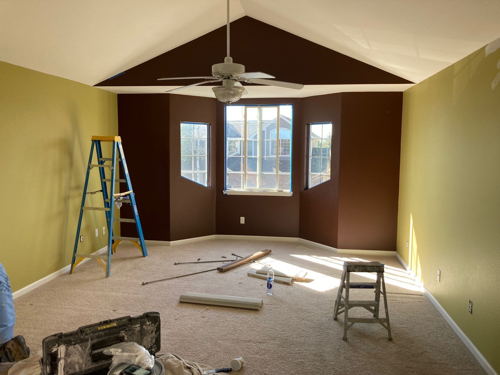 Room with brown accent wall, tan walls, ceiling fan, and ladder; under construction.