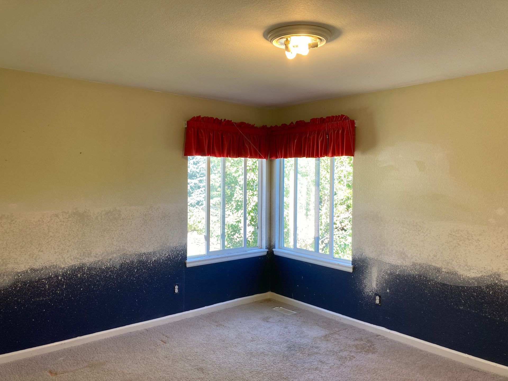 Empty room with corner window, blue and tan walls, red valance, ceiling light, and carpet.
