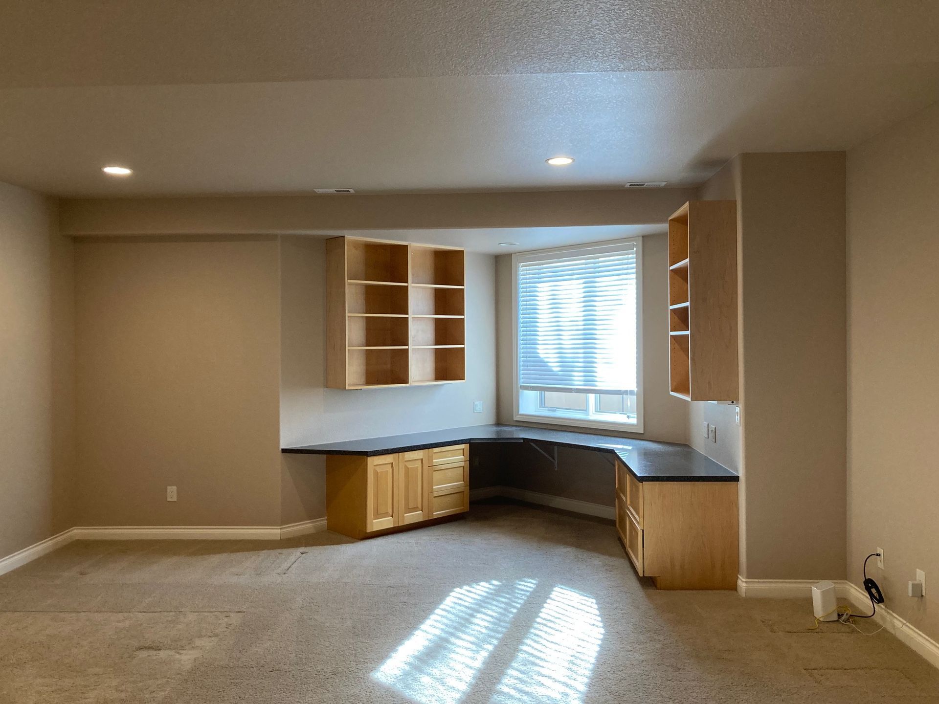Empty room with built-in desk, cabinets, and shelving around a bay window. Beige walls and carpet.