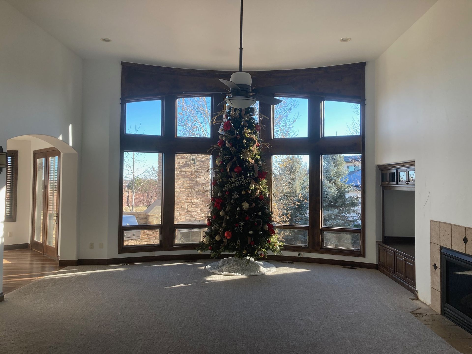 Large living room with a tall Christmas tree in front of a large window; sunlit space with a fireplace and wood trim.