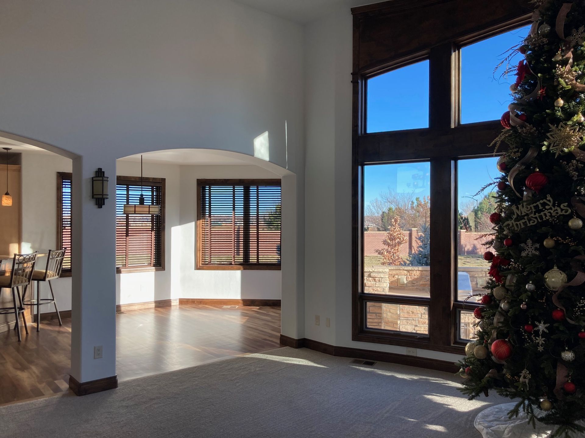 Spacious living room with Christmas tree, large windows, and an archway leading to dining area. Bright sunlight.