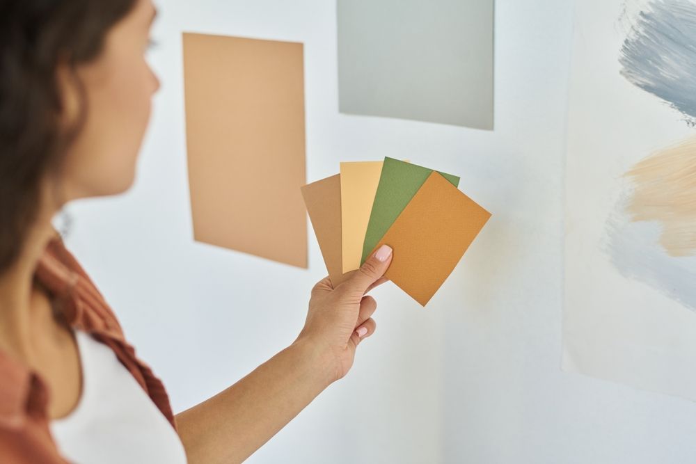 Woman comparing paint swatches to a wall, holding a variety of brown and green colors.