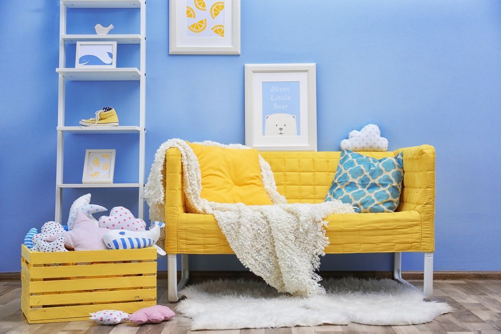 Yellow sofa with throw pillows and blanket in a blue room, with white shelving and artwork.