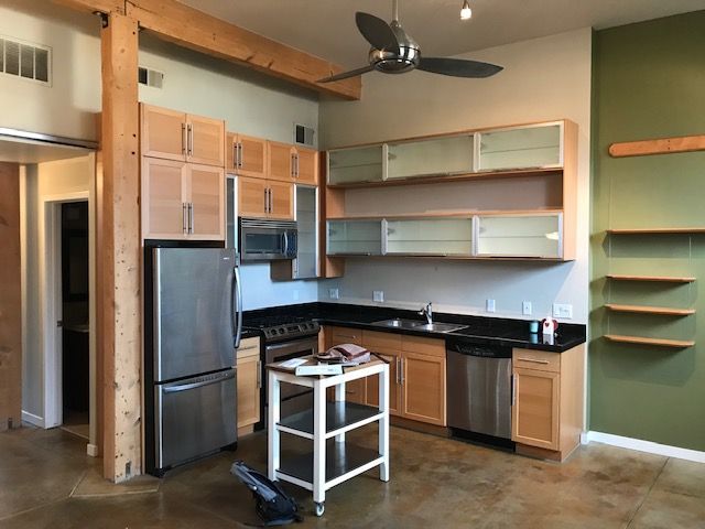 Modern kitchen with stainless steel appliances, light wood cabinets, and black countertops. Exposed beam and concrete floor.