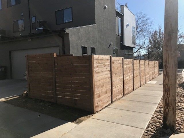 Brown wooden fence along a sidewalk next to a modern gray building.