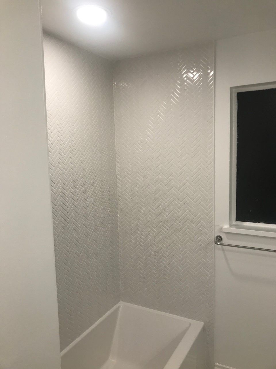 White tiled bathroom with a small tub. A window and light are visible.