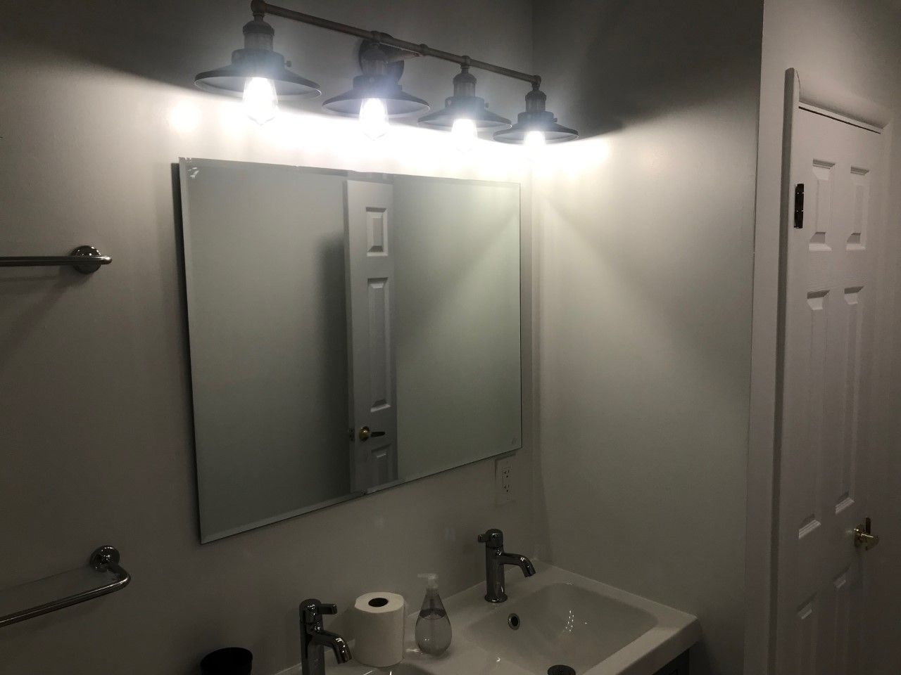 Bathroom with vanity, mirror, and four-bulb light fixture. White walls, chrome hardware, white sink, and closed door.